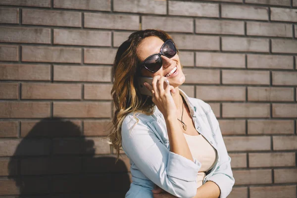 Cute cheerful female blogger dressed in trendy clothing spending free time outdoors and talking on mobile phone.Happy student in cool sunglasses communicating using gadget during summer walk outside