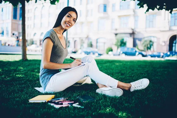 Capus の出費の時間のためのプロジェクトに取り組んでいるカジュアルな服装の若い女性を笑顔で中に芝生の上に座ってスケッチ ブックで図面を作る魅力的な陽気な女の子自由時間公園での夏の日 — ストック写真