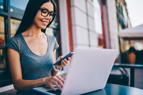 微笑的女性自由职业者在眼镜发送信息 而键盘在笔记本电脑有远程工作 Hipstergirl 安装在智能手机上使用蓝牙连接与上网本的应用 — 图库照片