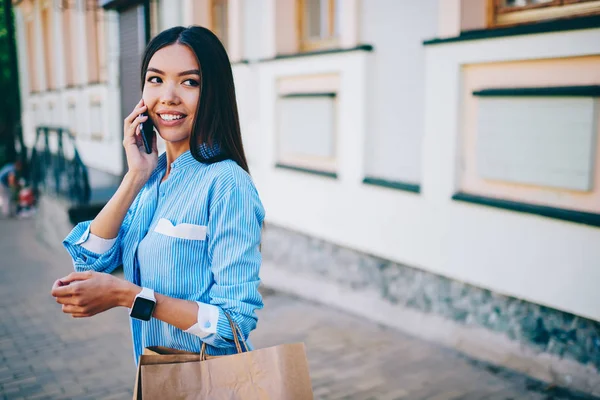 年轻漂亮的微笑时髦的女孩穿着蓝色的衬衫看着走 而打电话的智能手机持有购物袋在手 可爱的微笑的黑发妇女在城市花费闲暇时间 — 图库照片
