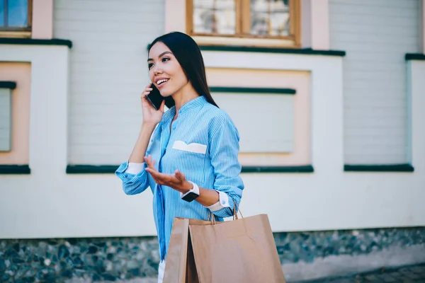 年轻迷人的亚洲女性微笑着通过智能手机进行手势 美丽的积极的时髦女孩走在城市设置的纸购物袋和手机设备交谈 — 图库照片