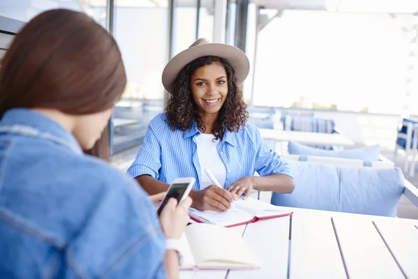 Portrait of cheerful dark skinned hipster girl writing in notepad during meeting with friend in cafe,happy young african american female student learning with colleague using smartphone for bloggin