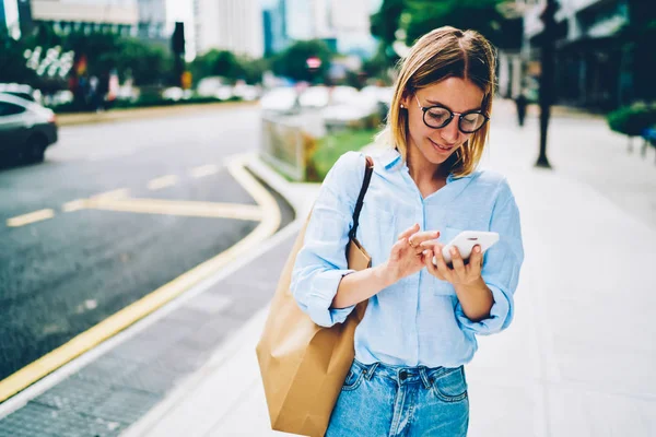 スマート フォン ダウンタウン内の移動のためのモバイル アプリケーションを使用して場所を検索金髪の女性旅行者の通りテキスト メッセージの上を歩くメガネやトレンディなカジュアルな服装の若い女性 — ストック写真