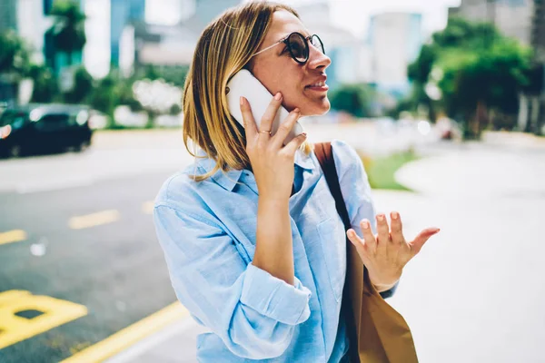 Smiling hipster girl in spectacles walking on street having phone conversation,positive female traveler enjoying city center using roaming connection for mobile call. Young woman exciting calling