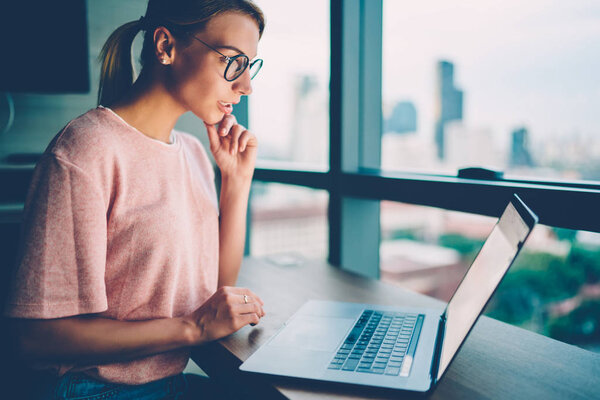 Потрясающая умная молодая женщина в электронных очках читает последние новости в социальных сетях на ноутбуке гаджете, проводя вечернее время дома.
