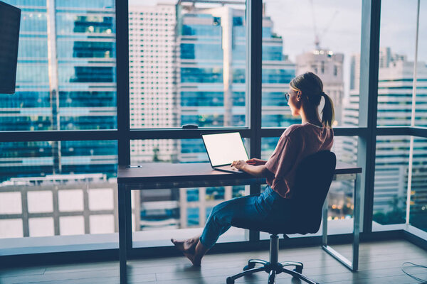 Обратный вид девушки хипстера в повседневной одежде в чате с друзьями в сети на современном компьютерном устройстве с пустым экраном для интернет-рекламы на сайте, сидящем в квартире
