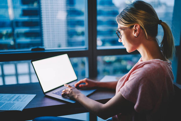 Молодая женщина в очках печатает текстовый пост для загрузки на интернет-сайт на нетбуке с пустым экраном для копирования. Женщина тщательно работает над курсом, сидя дома за компьютером ноутбука
