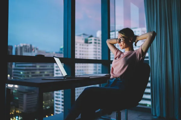 Attractive female freelancer resting after complete work in office on laptop with blank screen. Student enjoying recreation time watching movie on computer at home interior with evening city view