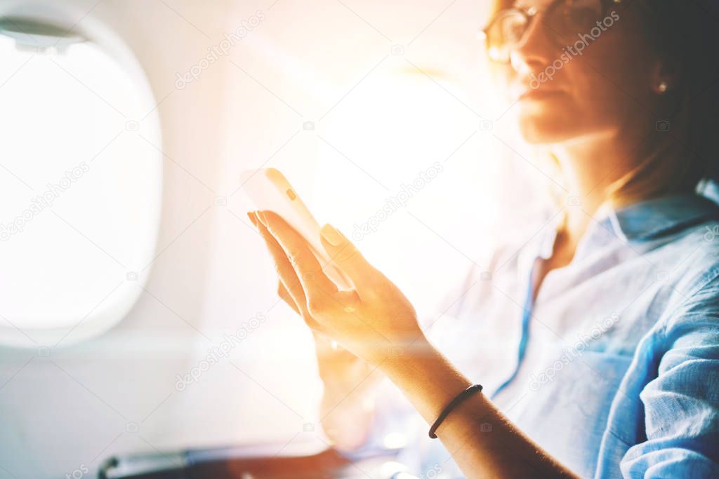 Close up of young caucasian female passenger of airplane using internet wireless connection on board enjoying comfortable service of first class. Woman browsing flight wifi network on smartphone