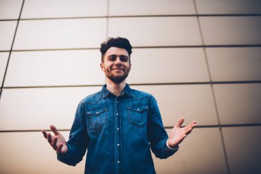 Kollarını açarak açık havada Selamlar, modaya uygun kot gömlek el hoş geldiniz işareti ve gülümseyen duvar arka plan üzerinde poz erkek hipster hazır duran pozitif yakışıklı sakallı adam portresi