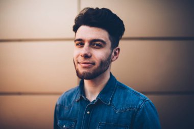 Kamera, yakışıklı hipster adam trendy mavi kot gömlek markası veya etiket için kopya alanı ile bakarak duvar arka plan üzerinde son moda saç modeli duran yakışıklı genç adam portresi