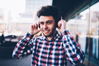 Genç delikanlı Caddesi üzerinde boş zaman serbest zaman sırasında zevk kulaklık ile gülümseyen hipster adam serin ekose gömlekli kentsel ayarları arka plan, fotoğraf makinesi ayakta bakarak gülen portresi