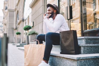 Trendy mutlu Afro-Amerikan erkek alıcı giymek başarılı alışveriş hakkında arkadaşıyla telefonda konuşurken, çanta ile merdivenlerde oturan işleci ile mobil konuşmayı adam neşeli koyu derili