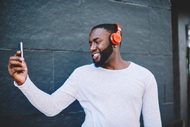 Erkek hipster koyu renkli selfie müzik dinleme sırasında yapma gülümseyen neşeli Afrikalı-Amerikalı adam modern araçlar smartphone kamerada fotoğraf çekimi ile açık havada vakit kulaklık