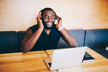Kapalı gözlerle kullanarak modern kulaklık ve laptop, pozitif afro Amerikalı adam radyo yayın yolu ile kullanma netbook üzerinde zevk çalma listesinden favori müzik dinlerken adam esmer gülümseyen
