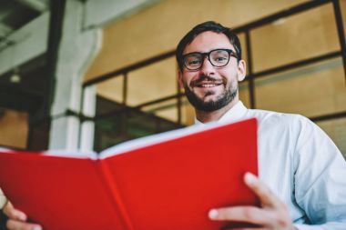 Yarım uzunluk portre pozitif yaratıcı çalışanın ofisinde oturup Not Defteri'nde muhasebe ile bazı iş yapma gömlek süre giymiş. Gülümseyen erkek kişi kapalı çalışırken kameraya bakıyor