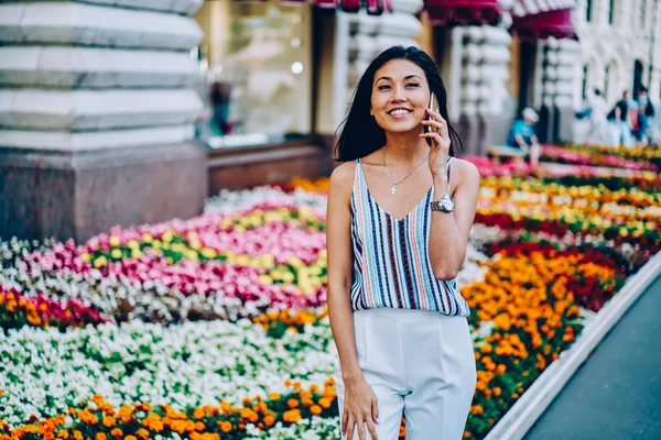 Cheerful female tourist satisfied with tariffs for calls in roaming talking with friend on mobile walking on street, positive asian girl in stylish apparel smiling during phone conversation