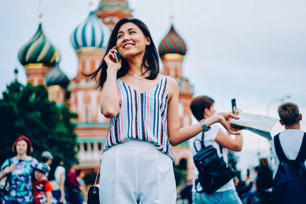 Cheerful female traveler satisfied with city tour in Moscow sharing emotions during mobile conversation, happy asian traveler using roaming connection for making phone calls during trip abroad