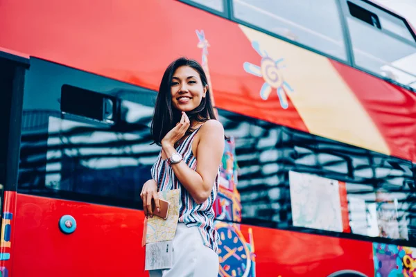 Portrait of attractive female traveler waiting for starting excursion on red sightseeing bus, happy young asian woman looking at camera planning to make city tour in Moscow city using transport