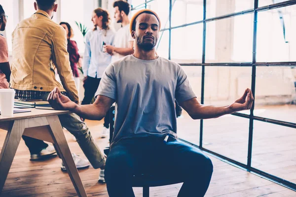 Afro american male member of working team sitting with closed eyes in lotus pose feeling peace breathing while colleague having discussion, skilled designer finding inspiration in meditation on work