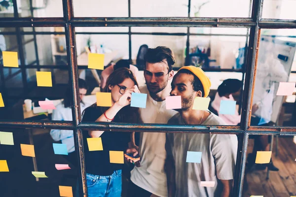 Male and female members of young crew collaborating near transparent wall with stickers pondering on strategy, skilled designers solving project problems together looking at memo cards with ideas