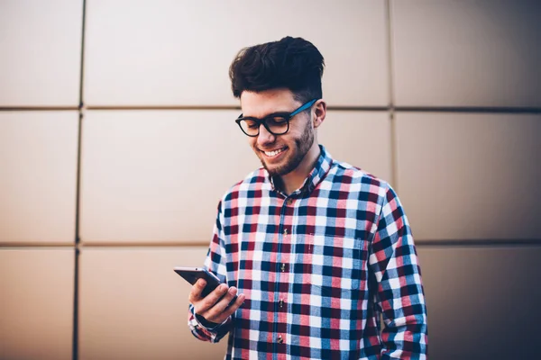 Cheerful hipster guy in plaid shirt  happy receiving message from friend on mobile phone, young male student in eyewear checking email on cellular excited with getting discount for sending SMS