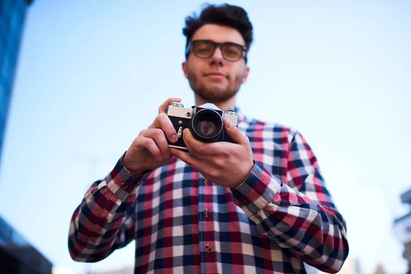 Selective focus on vintage camera in hands of hipster guy standing outdoors, young handsome traveler fond of photography taking pictures on old retro equipment during free time on city street