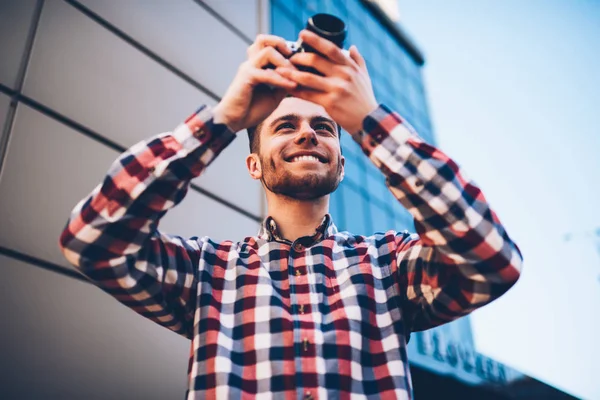 Smiling young skilled man taking photo standing on city street using classic vintage camera enjoying work, prosperous male photographer in plaid cool shirt making creative  images on urban settings