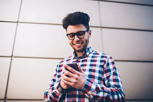 Cheerful male student in eyeglasses blogging in social networks via mobile chatting with followers, smiling hipster guy in plaid shirt receiving photos from friend on email while standing outdoors