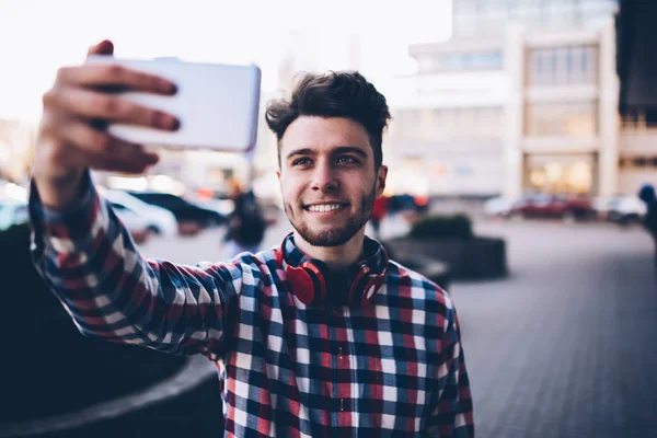 Cheerful male blogger in trendy outfit shooting video on smartphone walking on city street, smiling hipster guy making selfie application on mobile phone spending time time outdoors on urban settings