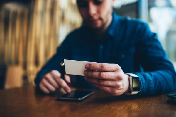 Selective focus on blank visit card in hands of hipster guy using email address for sending message via mobile, young man contact banking service online getting email from visit card for consultancy