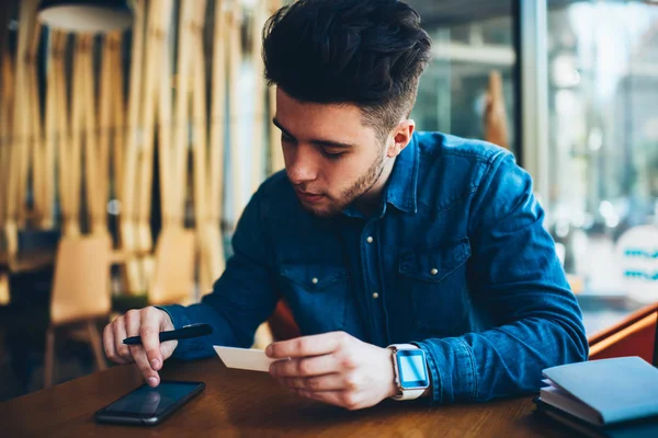 Pensive hipster guy searching website on mobile phone using address from card sitting in cafe,young man searching contacts of banking service for consultancy online holding visit card with information