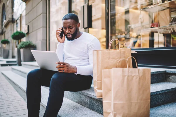 Pensive dark skinned guy talking on mobile with operator while making payment for purchase in web store via laptop,afric american man having mobile conversation about delivery getting bags with buying