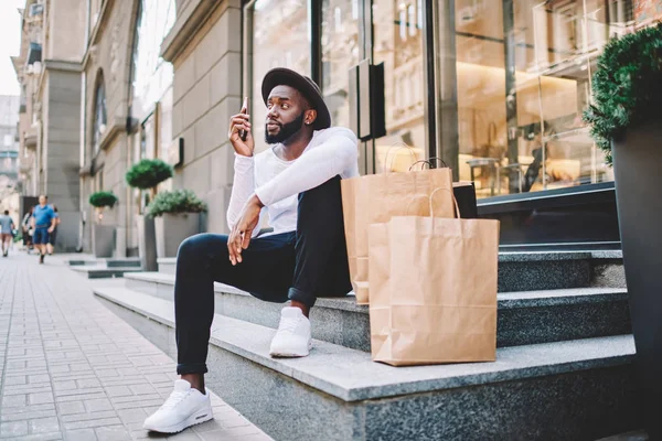 Serious african american male shopper making phone call for contact service operator waiting for delivery, young dark skinned man sitting on stairs of shop near paper bags with copy space for label