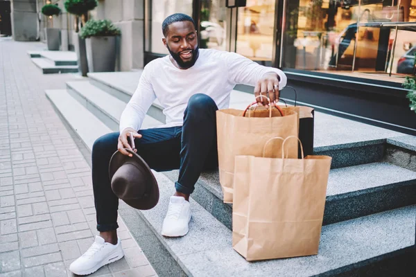 Smiling african american male shopper checking purchases in paper bags with coy space for brand name or label sitting on stairs, dark-skinned hipster guy resting outdoors after buying stuff on sales