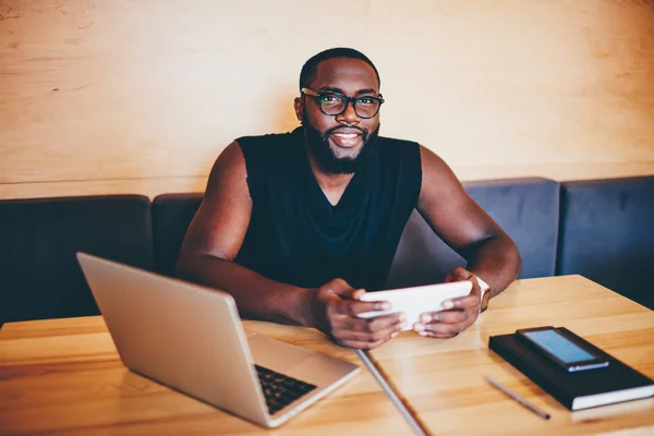 Portrait of cheerful bearded dark-skinned freelancer using modern technologies doing remote job in cafe, skilled afro american entrepreneur synchronizing files with  laptop using application on tablet