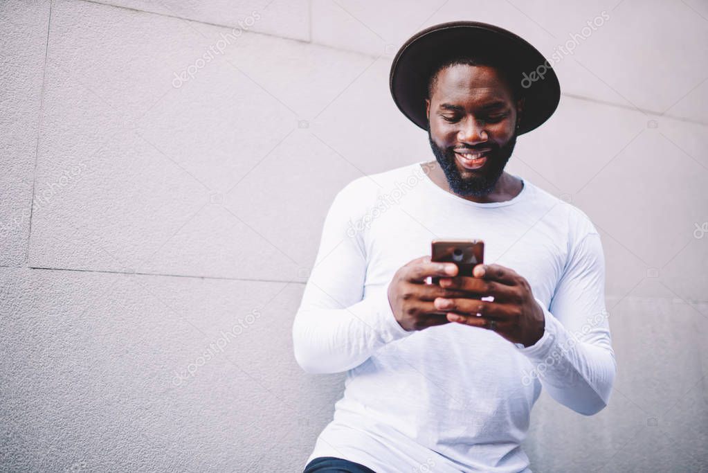 Trendy dressed African american hipster guy in feeling happy while chatting via application on mobile phone, dark-skinned man reading news from websites standing with smartphone near copy space area