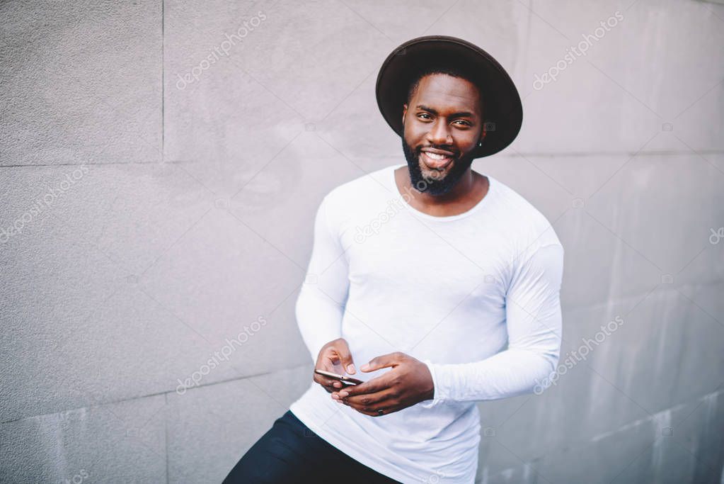 Portrait of cheerful afro american man posing while chatting in social network via cellphone standing on copy space background, smiling dark skinned hipster guy holding modern smartphone in hands