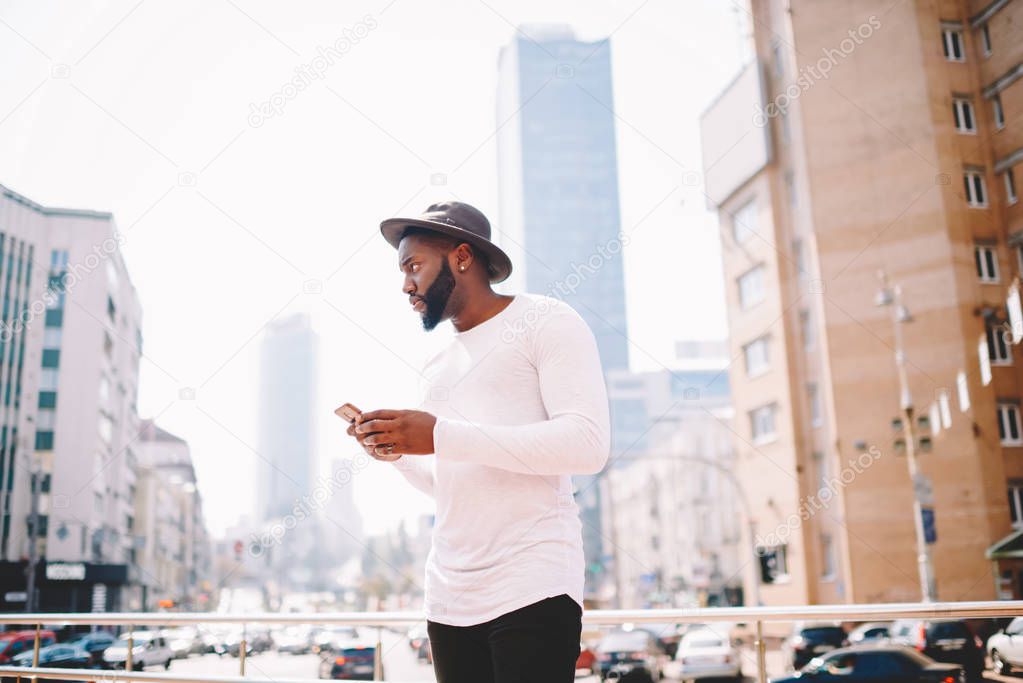 Serious afro american man in trendy outfit sending messages while standing on urban setting background, thoughtful dark skinned hipster guy in hat using application for navigating in megalopolis