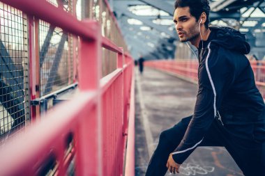 Motive fit adam kulaklık müzik mükellefi dinleme ve Koşudan önce ısınma yapıyor şık etkin elbise giymiş. Köprüde önce egzersiz germe yapıyor atletik erkek koşucu