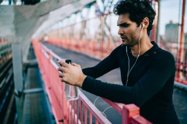 Ciddi uygun adam kulaklık şarkıları dinleme ve müzik uygulama ve kentsel ortamda spor çalışmadan sonra smartphone üzerinde yükleme aktif giyim giymiş. Köprüde elinde duran şişe ile sporcu