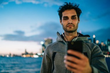 Smartphone aygıt üzerinde müzik uygulama yükleme ve açık havada akşam saat içinde kentsel ortamda ayakta modern kulaklık ses şarkıları dinleme spor ceket giymiş adam genç sakallı