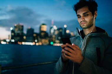 Uygulama yükleme adam genç sakallı ve ışık smartphone ekranında yansıtılır. Sakallı erkek blogger akşam kentsel ortamda hücresel ayakta bağlı earpones içinde müzik dinleme
