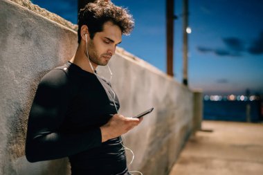Etkin giymek için müzik çalma listesinde seçtiğiniz elinde telefon ile kentsel ortamda eğitim ayakta yan görünüm atlet giymiş. Adam akşam Player'a bağlı kulaklık ses dinleme uygun