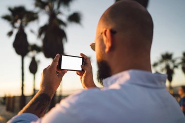 Erkek blogger fotoğraf çekmek için sahte ekranı ile smartphone kullanarak görünümünü sırt ve ağlarda, modern cep telefonu fotoğrafları yaz tatillerini akşamları sırasında yapma hipster adam pay