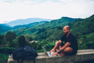Dizüstü bilgisayar aygıt açık havada tepede yeşil manzaralar ve mesafe işlemi için yüksek hızlı internet bağlantısı kullanılarak Dağları güzel manzara ile oturan klavye dalgın sakallı genç adam turizm