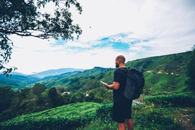 Erkek turist ile siyah sırt çantası holding smartphone online navigator ve uzak Asya yerlere sefer sırasında yüksek yeşil tepelerin ve manzara zevk ile. Uzun yürüyüşe çıkan kimse sırasında mountaints içinde yolculuk tutkusu