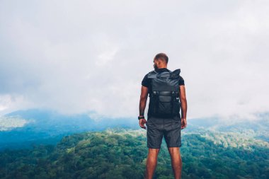 Rear view of exerienced male tourist with black backpack climbed to top of mountain and admiring amazing scenery of high hills with green natural environment under cloudy sky during expedition clipart