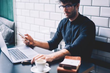 Kahve Dükkanı netbook aygıt üzerinde finansal hesap kontrol yetenekli erkek sahibi. İşadamı gözlük ve haber Web sitesinde dizüstü bilgisayar Internet bağlantısı okuma elinde smartphone