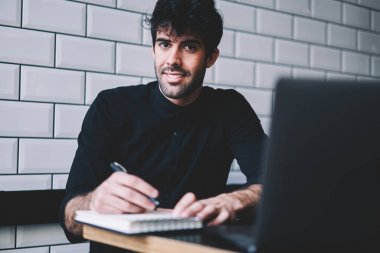 Sırasında e-öğrenme coworking dizüstü bilgisayar, kamera graffitti Not defterinizdeki notlar aşağı süre gülümseyen neşeli yakışıklı öğrenci portresi. Kayıt Defteri'nde belirterek olumlu genç adam